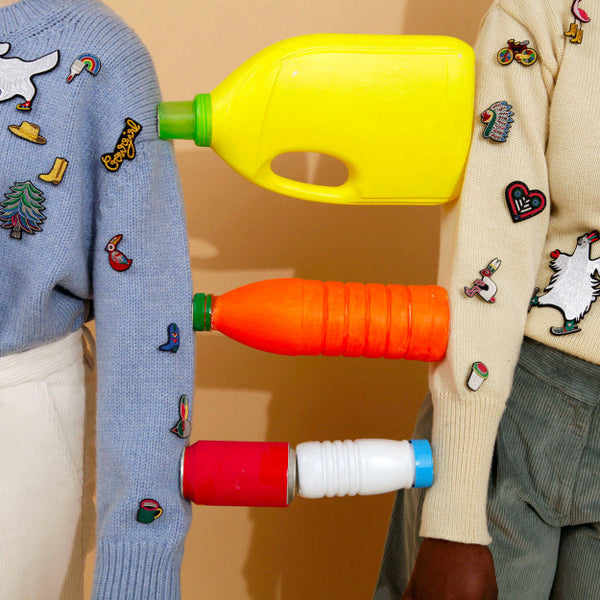 Two arms with a blue and cream jumper, decorated with hand embroidered colourful brooches holding bright bottles. Designs by Macon et Lesquoy, available to purchase at www.cuemars.com